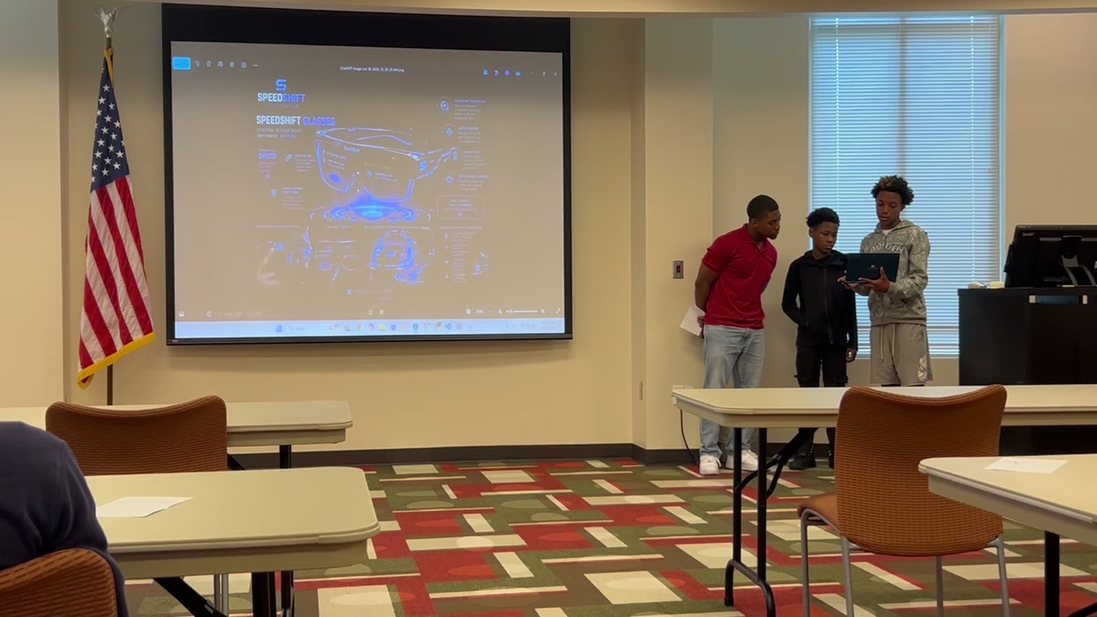 Three students at the front of the room presenting their battle bot design, with the bot schematic projected behind them